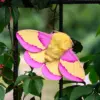 Handmade figurine Rosy Maple Moth on a fence amidst green foliage.